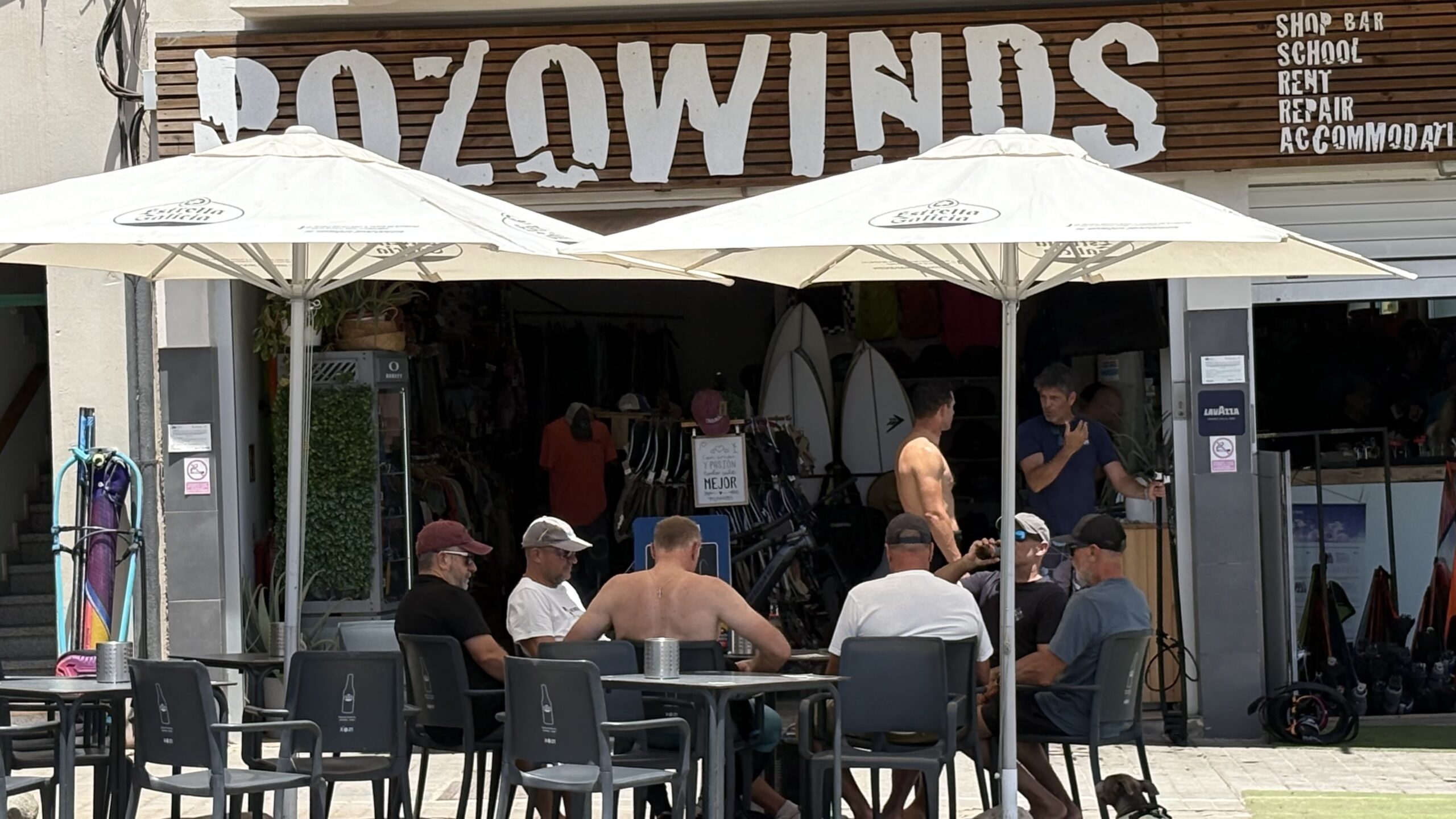 Menschen sitzen vor dem Surfshop POZOWINDS zwischen Surfboards in Pozo Izquierdo